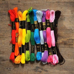 May include: A collection of colorful embroidery floss in shades of red, orange, yellow, green, blue, gray, purple, pink, and brown. The spools are arranged on a wooden surface, showcasing a variety of colors for crafting projects.