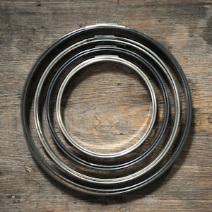 Puede incluir: Un conjunto de cuatro aros metálicos circulares encajados, cada uno con un pequeño mecanismo de tornillo ajustable en la parte superior. Los aros varían en diámetro, siendo el aro más interno de tono plateado y los tres externos de tono oscuro.