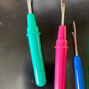May include: Three seam rippers with metal blades and plastic handles in bright green, hot pink, and blue. Each tool features a pointed metal fork with a small red safety ball at the tip. The green and pink rippers have matching plastic caps.