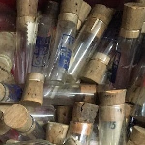 May include: A collection of vintage glass vials with cork stoppers. Some vials have labels with blue text. The vials are of varying sizes and shapes, and some contain coloured substances. The corks are aged and show signs of wear.