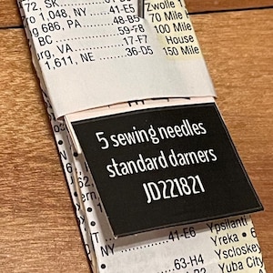 Peut inclure: Un paquet de 5 aiguilles à coudre étiquetées standard darners JD221821. L'emballage présente une couverture en papier blanc avec du texte noir listant divers lieux et codes, et une étiquette rectangulaire noire avec du texte blanc.