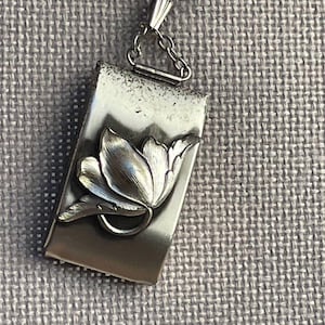 May include: A rectangular silver-toned metal pendant featuring a raised, brushed-finish floral motif on its front surface. The pendant is attached to a small metal jump ring and a short segment of a delicate silver-toned chain at the top edge.