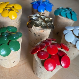 May include: A collection of six cork bottle stoppers, each topped with a cluster of colorful, round, metal accents. The colors include yellow, blue, teal, gold, green, red, and white. The stoppers are arranged on a light brown surface.