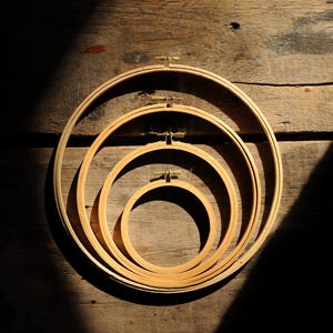 Puede incluir: Un conjunto de cuatro bastidores de bordado circulares anidados hechos de madera de color claro. Cada bastidor cuenta con un pequeño mecanismo de tornillo de ajuste de metal en la parte superior para fijar las anillas interiores y exteriores.