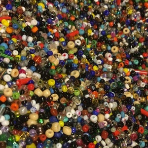 May include: A dense collection of small, multi-coloured glass seed beads in various shapes and sizes. The assortment includes opaque, translucent, and metallic finishes in shades of red, blue, green, yellow, black, white, and tan.