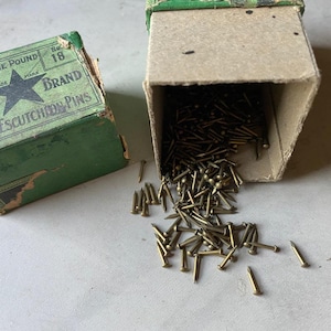 Pode incluir: Uma caixa de cartão vintage com etiqueta de papel verde e logótipo de estrela preta, rotulada como One Pound No. 18 Star Brand Escutcheon Pins. A caixa está aberta e derrama uma grande quantidade de pequenos pregos cor de latão.