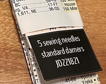 Puede incluir: Un paquete de papel con 5 agujas de coser, específicamente agujas de zurcir estándar, con el texto JD221821 impreso en una etiqueta negra. El empaque presenta un fondo blanco con texto negro que enumera varias ubicaciones y códigos.