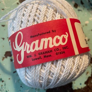 May include: A ball of white twisted string wrapped in a red paper label. The label features the white text Gramco, Manufactured by, and WM. J. GRAHAM CO., INC. Lowell, Mass., 01854. The string has a thick, braided texture and is wound into a round shape.