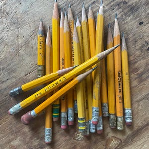 Puede incluir: Una colección de lápices de madera amarillos con puntas de grafito y virolas de metal. Varios lápices presentan texto que incluye 'CAMPUS No 2' y 'Land O'Lakes, Inc. No 2'. Los lápices están dispuestos en un grupo, mostrando longitudes y desgaste variables en la madera.