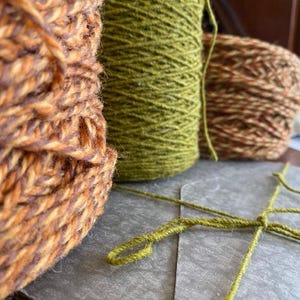 Könnte beinhalten: Drei Garnknäuel in verschiedenen Texturen und Farben. Eines ist eine dicke, melierte Mischung aus Orange und Braun, ein weiteres ist einfarbig olivgrün und das dritte ist eine mehrfarbige Mischung aus Braun und Orange. Ein olivgrüner Faden ist verknotet.