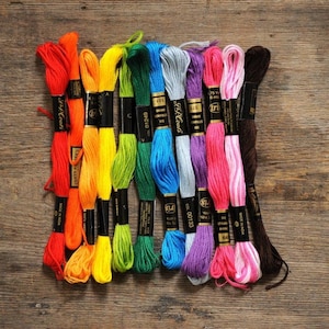 May include: A selection of embroidery threads in a spectrum of colours, including red, orange, yellow, green, blue, grey, purple, pink, and brown. The spools are displayed on a wooden surface, offering a range of hues for craftwork.