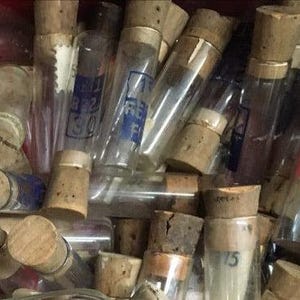 Könnte beinhalten: Eine Ansammlung von zylindrischen Fläschchen aus klarem Glas, die jeweils mit einem hellbraunen Korkstopfen verschlossen sind. Mehrere Fläschchen weisen aufgedruckte blaue oder rote Texte und numerische Markierungen auf der Oberfläche auf.