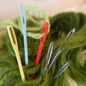 May include: Close-up of several sewing needles in various colors, including yellow, blue, and silver, amidst a tangle of green yarn. A red needle is threaded with light green yarn. The image highlights the tools used for sewing and crafting.
