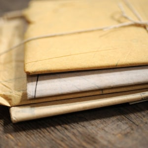 May include: A stack of three rectangular paper envelopes in shades of tan and light gray, secured together with thin white twine. The top envelope is a light tan color, while the middle one is a pale gray, and the bottom is a darker tan.