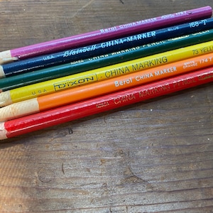 Può includere: Una collezione di cinque matite a cera tipo pennarello per porcellana nei colori viola, blu, verde, giallo, arancione e rosso. Ogni matita presenta una punta in carta a strappo e testo stampato che include marchi come Berol, Blaisdell e Dixon con etichette di colore.