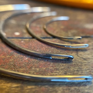 May include: Close-up of several curved needles with eyelets, arranged on a rustic, weathered wooden surface. The needles are made of shiny, silver-colored metal. The background is out of focus, highlighting the needles' details.