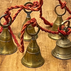 May include: Five brass bells of varying sizes are connected by a single, continuous red twisted cord. Each bell features a circular loop at the top for suspension. The bells have a metallic gold finish, and one bell displays the number 20 engraved.