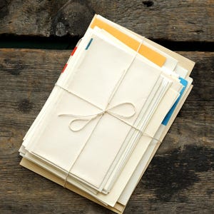 Könnte beinhalten: Ein Stapel cremefarbener Schreibwaren, mit Bindfaden zusammengebunden, liegt auf einer verwitterten Holzoberfläche. Der Stapel enthält Umschläge und Papier in verschiedenen Größen, mit Andeutungen von orangefarbenen, blauen und roten Kanten. Der Bindfaden ist zu einer sauberen Schleife gebunden.