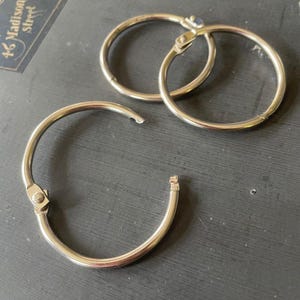 May include: Three silver-colored metal loose-leaf binder rings. Two of the rings are closed in a circular shape, while one ring is open, showing the hinged mechanism. The rings have a smooth, polished finish and a simple, functional design.