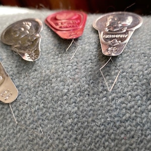 May include: A collection of vintage silver and red metal sewing needle threaders. The threaders are in various shapes, including a shield, a diamond, and a coin-like design. They are resting on a textured, light blue fabric.