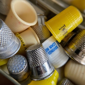 May include: A close-up of a collection of thimbles in a variety of materials and colours. There are silver metal, yellow plastic, and cream plastic thimbles. Some have dimpled surfaces, others are smooth. One has the number 9. The text "SAVINGS LOAN ASS" is visible on one.