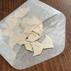 Op de afbeelding: Een verzameling beige papieren hoekbeschermers, sommige met reliëf geometrische ontwerpen, zijn gedeeltelijk zichtbaar in een doorschijnende envelop. De envelop is open en onthult de inhoud. De achtergrond is een houten oppervlak.