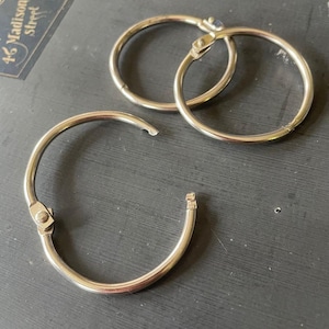 May include: Three silver binder rings are displayed against a dark background. The rings are open, showing their hinged design. The metal has a shiny finish, reflecting light. The rings are likely used for organizing papers or other materials.