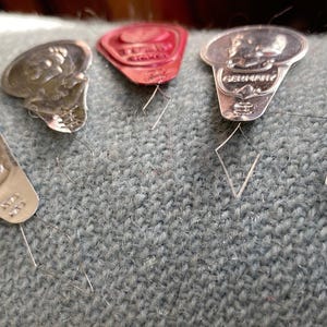 May include: Five metal needle threaders arranged in a row. Four are silver-toned with embossed designs and the word GERMANY, while one is red with a central embossed detail. Each features a thin, diamond-shaped wire loop for threading needles.