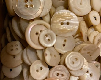 Puede incluir: Una colección de botones redondos variados en diversos tonos de blanco roto, crema y beige nacarado. Los botones presentan diferentes tamaños y configuraciones de agujeros, incluyendo diseños de dos y cuatro agujeros, con superficies lisas e iridiscentes.