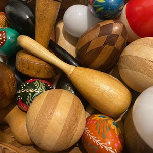 Puede incluir: Una colección de objetos de madera, que incluye esferas, formas de huevo y un mortero y maja. Las esferas varían en color y diseño, algunas con rayas, patrones y detalles pintados. Los objetos están dispuestos en una cesta tejida.