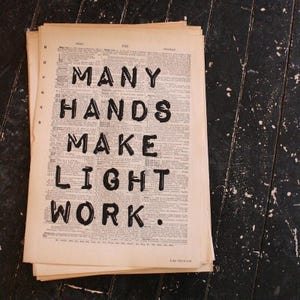 Puede incluir: Una pila de páginas de diccionario antiguas con el texto impreso en negro MANY HANDS MAKE LIGHT WORK centrado en la hoja superior. El papel es de color blanco roto con columnas visibles de definiciones y el nombre John Heywood al pie.