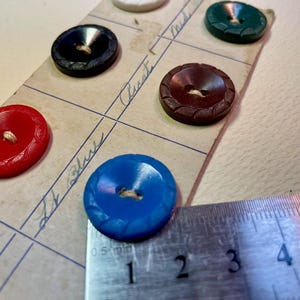 May include: A collection of five round buttons in red, blue, black, brown, and green, each featuring a textured edge and two center holes. A metal ruler is positioned next to the blue button, showing a diameter of approximately 1.5 centimeters.