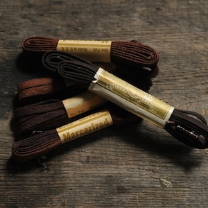 Puede incluir: Una colección de cordones planos de color marrón oscuro agrupados con bandas de papel color canela. Las bandas presentan el texto "Mercerized" y "One Pair Made in U.S.A." junto con un logotipo. Los cordones están tejidos con textura visible y tienen herretes.
