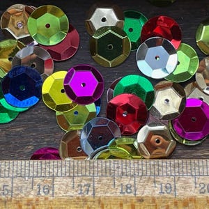 May include: A collection of multicolored, faceted, circular sequins with central holes, scattered above a wooden ruler showing measurements in inches. The sequins feature metallic finishes in shades of red, green, blue, yellow, purple, and silver.