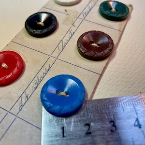 Puede incluir: Una colección de botones vintage en varios colores, incluyendo rojo, azul, negro y marrón, están dispuestos en una cuadrícula. Una regla metálica mide el diámetro de un botón azul, aproximadamente 1,9 cm. Los botones tienen un diseño texturizado y están sujetos a una tarjeta.