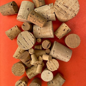 May include: A collection of various-sized corks, some with visible text, arranged on a red surface. The corks are primarily cylindrical, with a natural, light brown color and visible grain patterns. Some corks are cut at an angle.