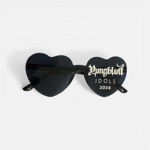 May include: Black heart-shaped sunglasses with the text "Yungblud IDOLS 2026" in white script. The sunglasses have a sleek, modern design, perfect for a bold fashion statement. The lenses are dark, offering a stylish look.