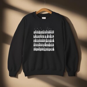 Könnte beinhalten: Ein schwarzes Langarm-Sweatshirt mit Rundhalsausschnitt und einem weißen Grafikdruck, der mehrere Reihen stilisierter immergrüner Bäume und kleine Vögel über der Waldlinie zeigt, präsentiert auf einem Holzkleiderbügel.