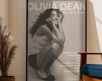 May include: A monochrome poster with the text "OLIVIA DEAN The art of loving" and a photograph of a woman. A blue rug with a decorative border is on a wooden floor. A blue armchair and a potted plant are also visible.