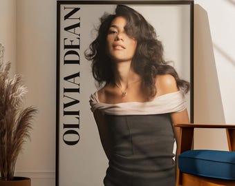 May include: A framed poster with the name "OLIVIA DEAN" in white lettering. The poster is leaning against a white wall, with a blue rug and a blue chair in the foreground. A potted plant and a window are also visible.