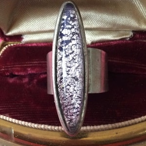 May include: A silver-toned ring featuring a long, vertical, marquise-shaped setting with a textured, shimmering purple and silver inlay. The ring is displayed inside a vintage-style, hinged jewelry box lined with dark red velvet and cream satin.