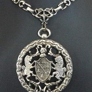 May include: Silver-coloured pendant necklace with a large circular pendant displaying a heraldic design. The pendant features a shield with two lions, surrounded by intricate detailing. The chain is a chunky, linked design.
