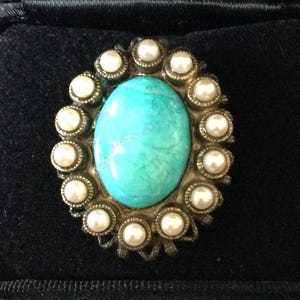 May include: Oval turquoise-colored stone brooch surrounded by small, white faux pearls. The brooch is set in a gold-toned metal frame and rests in a black velvet jewelry box. A vintage-style accessory.