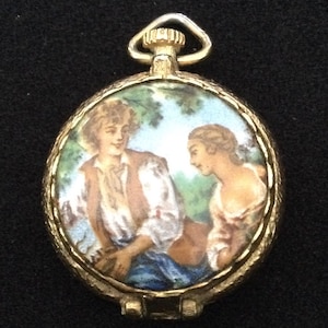 May include: A gold-toned, round pendant with a hinged lid. The pendant features a painted scene of two people in a garden setting. The background is a light blue sky with green trees. The pendant has a small loop at the top.