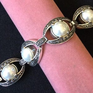 Peut inclure: Un bracelet argent&eacute; orn&eacute; de grandes perles blanches rondes, serties dans des maillons ovales d&eacute;coratifs. Le bracelet pr&eacute;sente un motif orn&eacute; et une fermeture &agrave; fermoir. Le bijou est pr&eacute;sent&eacute; sur une surface rose cylindrique sur fond noir.