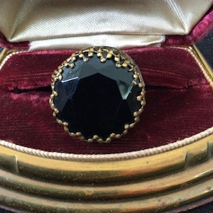 May include: A vintage black gemstone ring with a faceted cut, set in a gold-toned filigree setting. The ring is displayed in a velvet-lined jewelry box with gold trim. The box is open, revealing the ring.