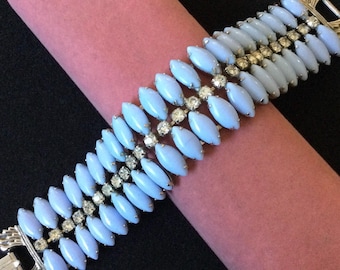 May include: A vintage bracelet featuring light blue oval-shaped beads and sparkling rhinestones. The bracelet has a silver-tone metal frame and clasp. The beads are arranged in rows, creating a layered effect. The bracelet is displayed on a pink velvet surface.