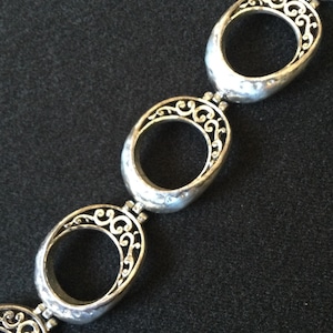 Może przedstawiać: Srebrna bransoletka z owalnymi ogniwami i skomplikowanymi ażurowymi wzorami filigranowymi. Bransoletka składa się z serii połączonych owalnych ogniw, z kt&oacute;rych każde ma szczeg&oacute;łowy, wirujący wz&oacute;r w owalnej ramie. Metal ma lekko młotkowaną fakturę.