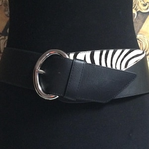 May include: A wide black belt with a silver-colored buckle. The belt features a section with a zebra print pattern in black and white. The belt is worn around the waist.