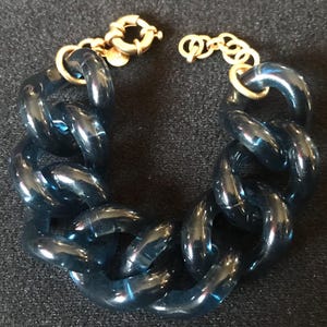 May include: A chunky chain bracelet with large, translucent blue links. The bracelet features gold-colored clasps and connectors. The links are rounded and have a glossy finish, creating a bold and stylish accessory.